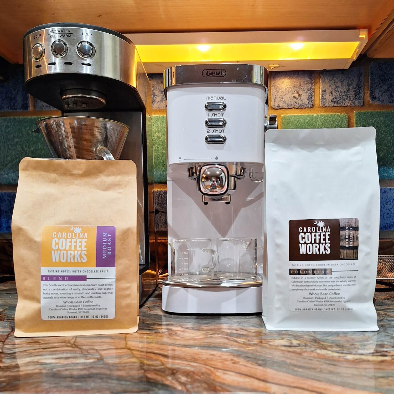 Two bags of carolina coffee works, medium roast and bourbon infused flavored coffee, on a kitchen counter with a coffee machine and other coffee appliances.