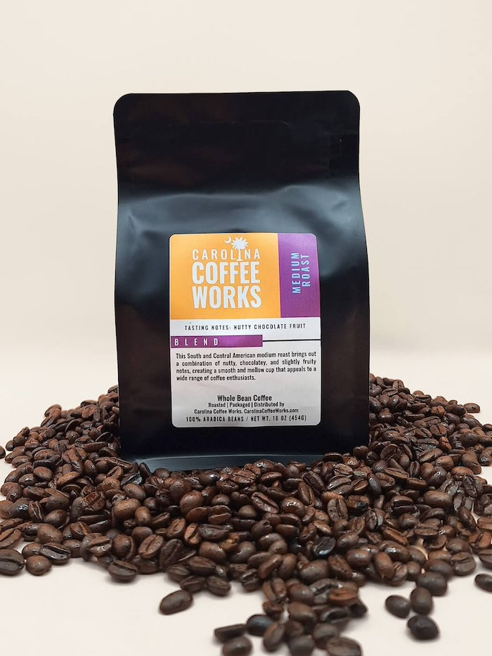 A bag of black medium roast coffee by Carolina Coffee Works standing on top of a pile of roasted coffee beans.