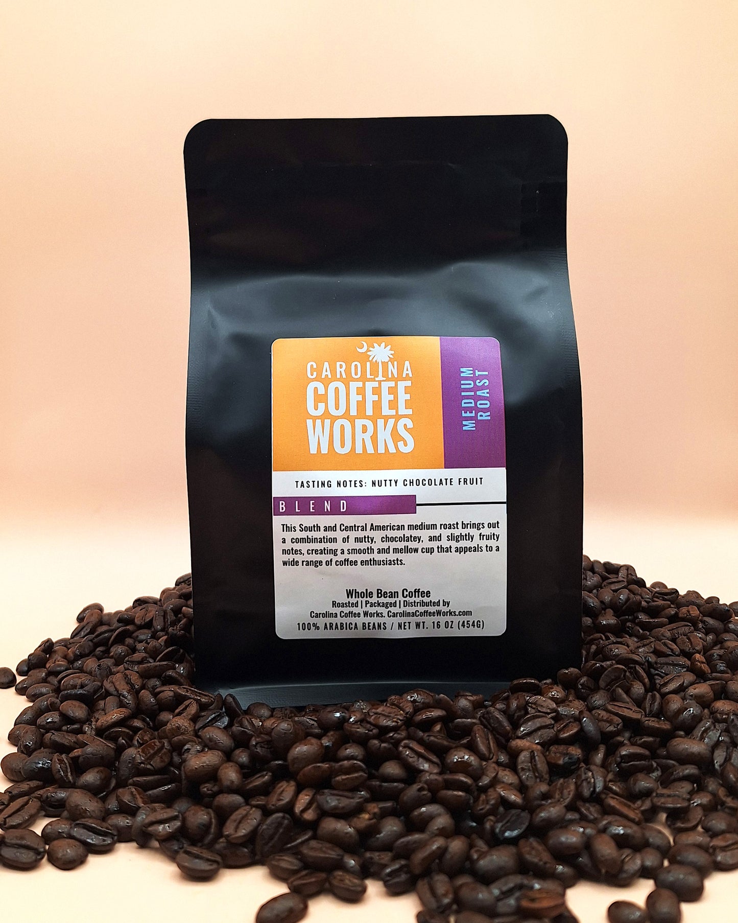 A black bag of Carolina Coffee Works Medium Roast Coffee placed on top of a stack of coffee beans against a tan background. 